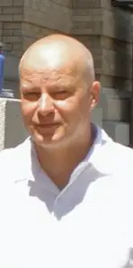Man with a shaved head wearing a white shirt outdoors.